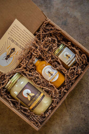 Three jars of honey in a box with bee-themed labels on a textured surface.